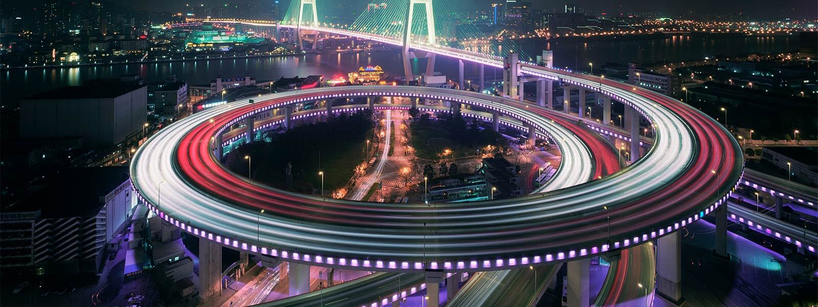 Fotografia em área urbana de uma ponte que atravessa um rio e possuí um viaduto circular iluminado