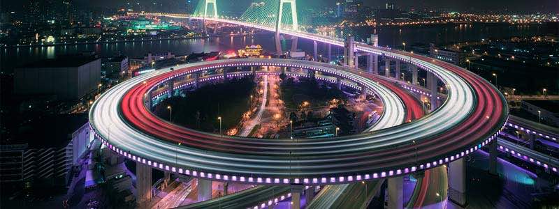 Fotografia em área urbana de uma ponte que atravessa um rio e possuí um viaduto circular iluminado