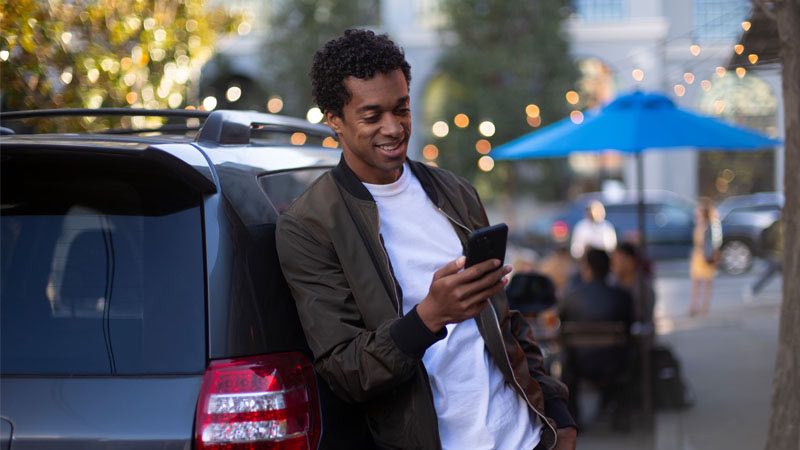 Homem encostado em um carro enquanto usa seu smartphone em local público