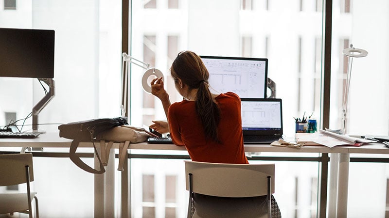 A woman sitting at a desk in front of a laptop and monitor with high-rise view.