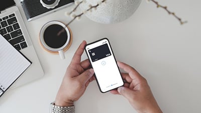 Person holding a smartphone displaying a Visa card that is powered by checkout.com in a digital wallet app, with a laptop, notebook, coffee cup, and camera on a white desk in the background.