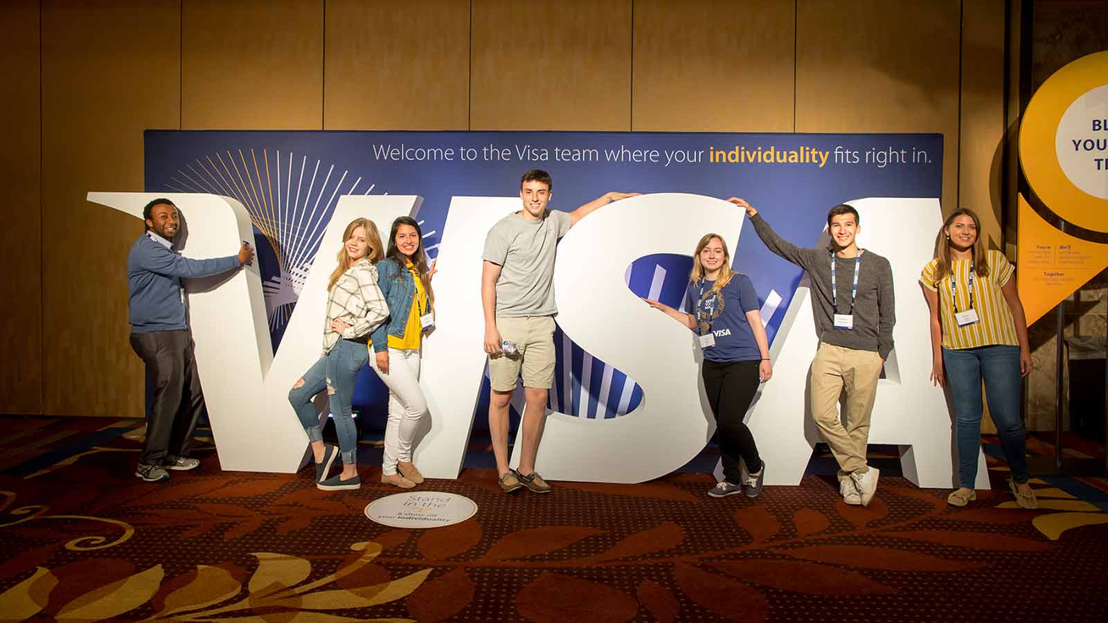 Group of seven young Visa employees next to large Visa logo letters