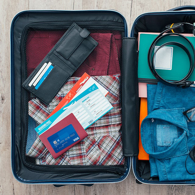 Open suitcase packed for travel with clothing, personal electronics, camera, wallet, airline tickets, passport and Visa card.