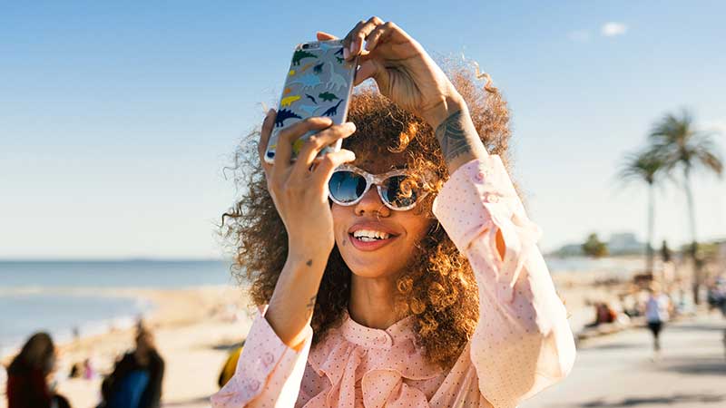 A woman takes a photo on her phone against the sunny coastline