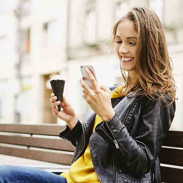 Mujer sentada en un banco mira su celular y come helado
