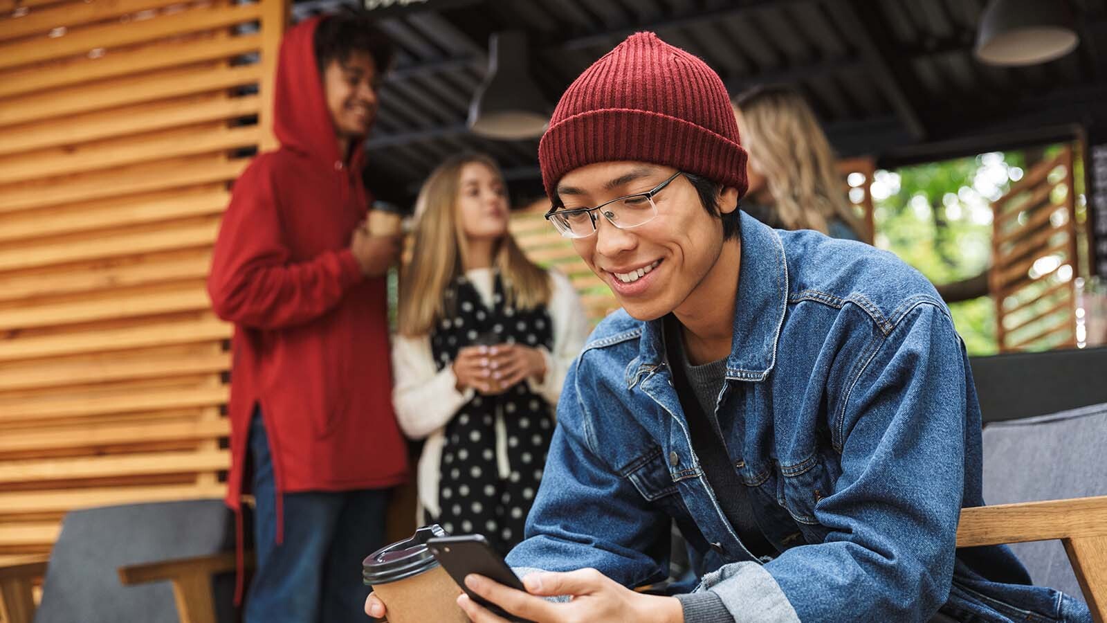 Imagem exibe jovem rapaz sorridente, sentado bebendo café e usando seu smartphone.