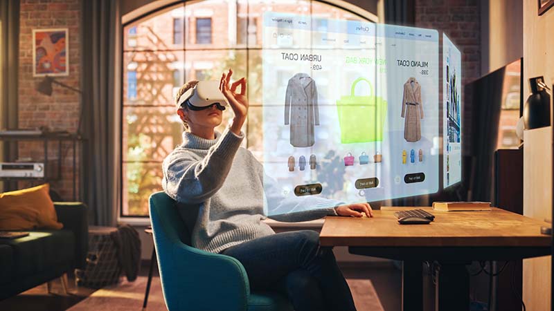 Imagem exibe mulher sentada com óculos de realidade virtual com holograma a sua frente, navegando em loja virtual de roupas. 