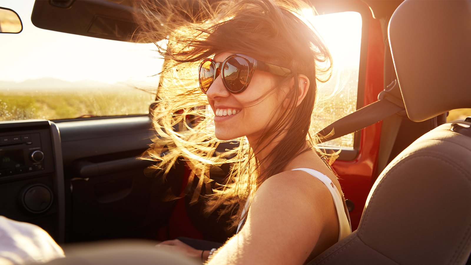 Mulher sorrindo dentro de carro, com óculos de sol e cabelos esvoaçando