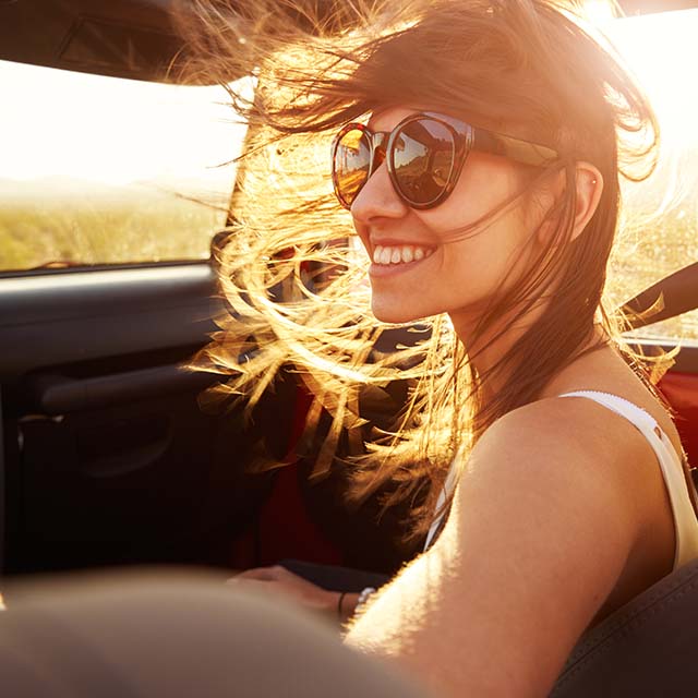 Mulher sorrindo dentro de carro, com óculos de sol e cabelos esvoaçando