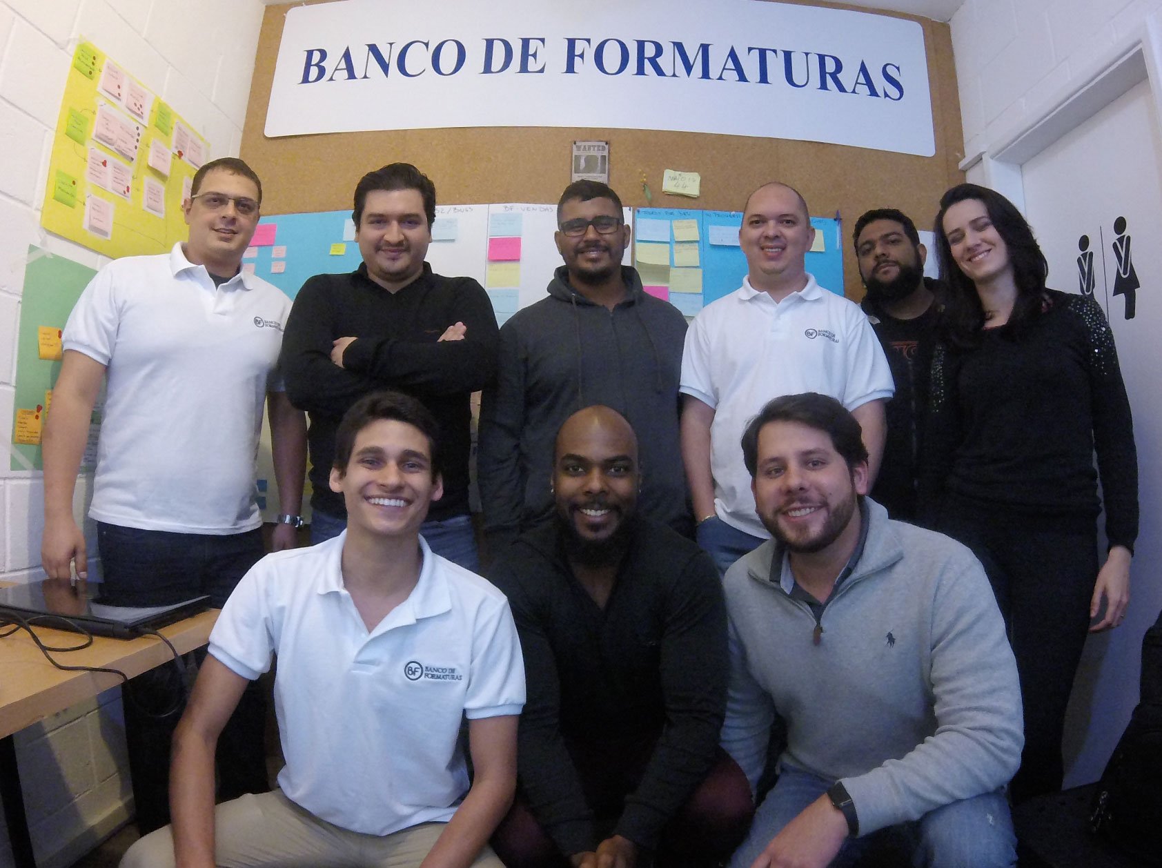 Grupod e homens posando para fotografia abaixo de uma placa onde se lê BANCO DE FORMATURAS