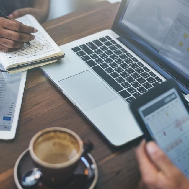 Notebook, iPad e xícara de café já vazia sobre uma mesa de madeira escura. No canto direito inferior desponta uma mão com celular, na tela se vê uma agenda com marcações. No canto superior esquerdo, mãos fazendo anotações em caderno