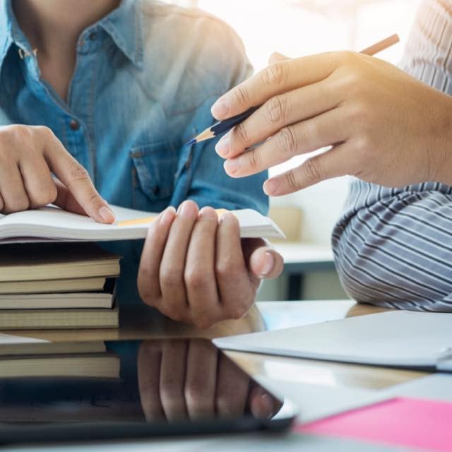 Duas pessoas estudam sobre uma mesa. Há vários cadernos empilhados, além de um iPad e um livro. As pessoas gesticulam