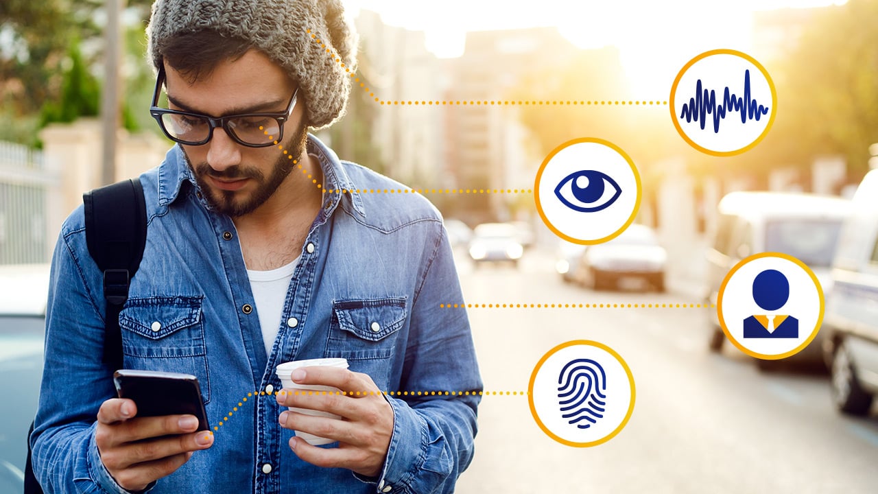 Homem de jaqueta jeans e gorro observando o celular com um copo pequeno de café na outra mão. Está conectado a símbolos digitais de biometria, olho e voz