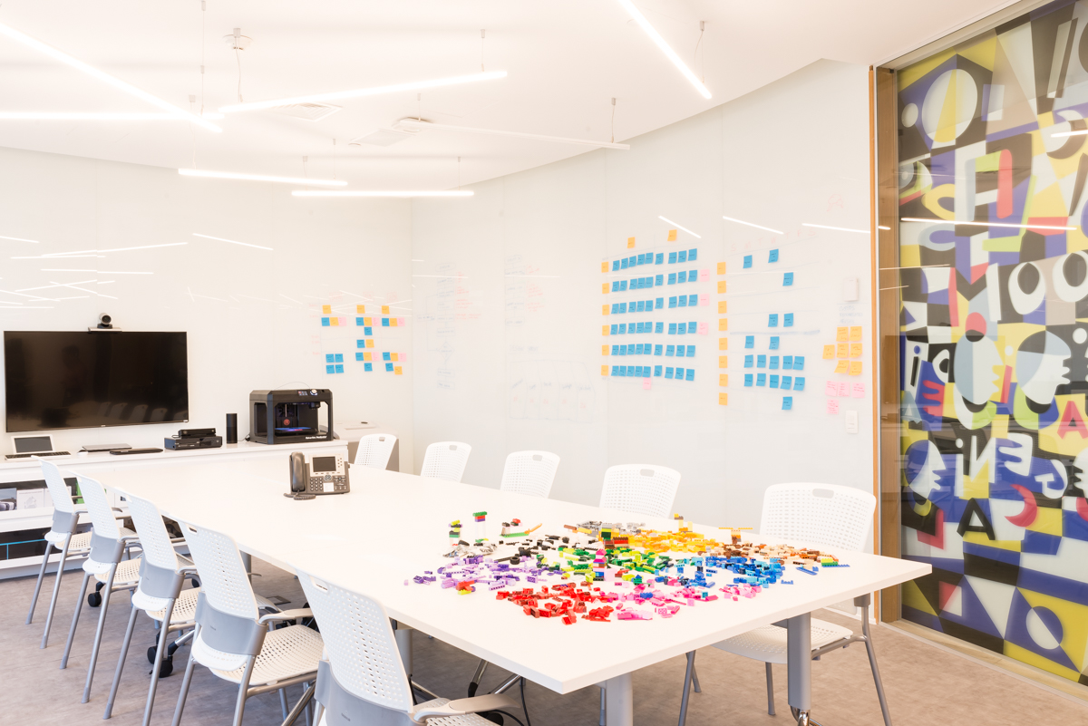 Sala de escritório criativo, com uma mesa branca retangular longa e cadeiras brancas dispostas ao longo dela. Na cabeceira da mesa, alguns equipamentos e um monitor com câmera. Há papéis coloridos em cima da mesa e post-its coloridos na parede direita, ao lado de um painel com pintura abstrata