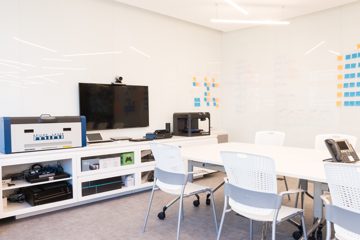 Sala de escritório criativo, com uma mesa branca retangular longa e cadeiras brancas dispostas ao longo dela. Na ponta da mesa, alguns equipamentos e um monitor com câmera