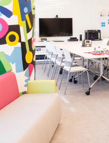 Visa Brasil Innovation Studio. À esquerda uma parede convexa com pintura abstrata em cores vibrantes e um sofá em tons de rosa e amarelo claros. No centro e na direita, uma mesa longa de escritório com cadeiras e uma TV.