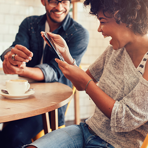 Mulher gargalhando com smartphone na mão enquanto um homem sorri para ela, os dois estão sentados num café
