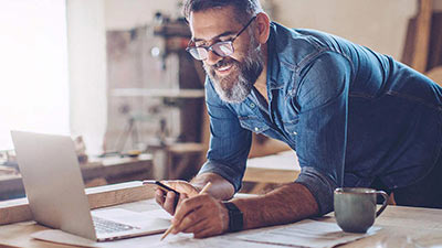 Homem de camiseta jeans debruçado sobre a mesa de trabalho. Faz anotações com lápis enquanto mexe no celular e sorri. Notebook e xícara sobre a mesa