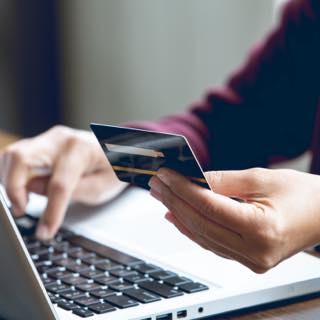 Pessoa fazendo pagamento online. Com uma mão segura o cartão e com a outra digita no notebook.