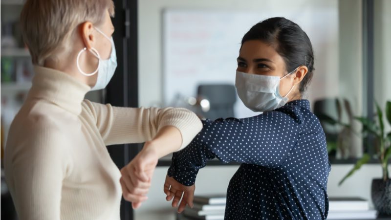 Duas mulheres utilizando máscaras de proteção sobre nariz e boca, cumprimentando-se com toque de cotovelo