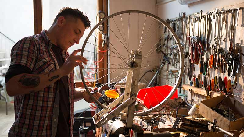 Homem tatuado observando a roda de uma bicicleta dentro da oficina