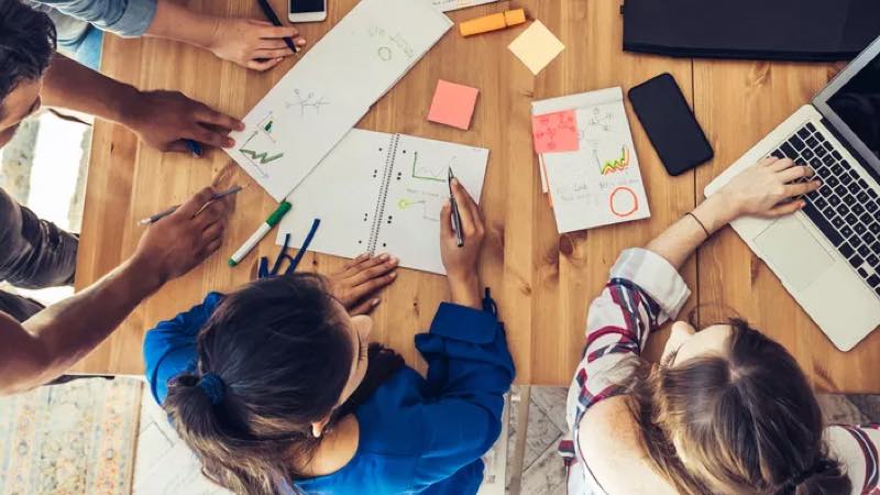 Vista do alto de pessoas trabalhando com notebook e agendas, analisando gráficos e conversando