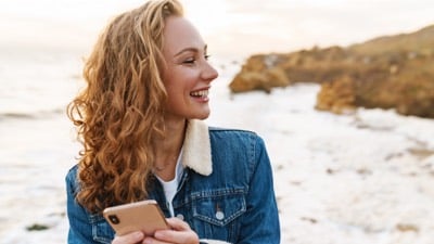 Imagem exibe mulher segurando celular enquanto sorri e olha pro lado