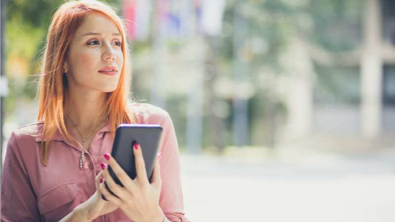 Mulher de cabelo alaranjado com um smartphone na mão observando algo na linha do horizonte