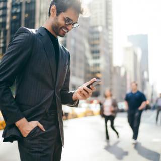 Homem com barba, de terno e camisa interna de cores escuras, usa óculos de grau. Está sorrindo e observando o celular na mão