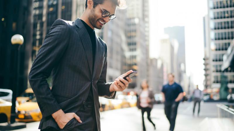 Homem com barba, de terno e camisa interna de cores escuras, usa óculos de grau. Está sorrindo e observando o celular na mão