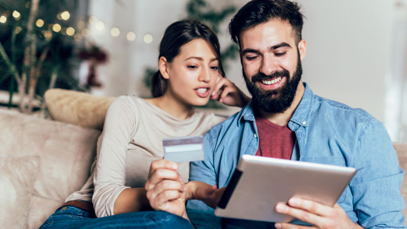 Imagem de um casal realizando compras online com um tablet e um cartão de crédito na mão