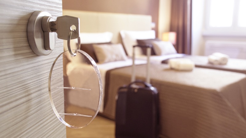 Imagem de um quarto de hotel com a porta aberta e uma mala ao lado da cama