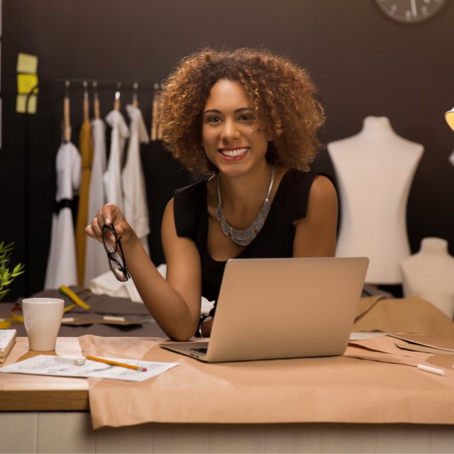 Imagem exibe mulher sorrindo em frente ao computador e segurando óculos