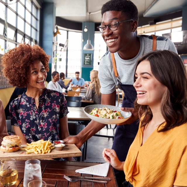 imagem exibe garçom sorrindo servindo dois pratos a uma mesa de quatro mulheres sorrindo