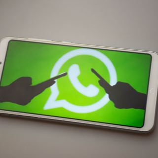 Tela de smartphone com símbolo do WhatsApp e silhuetas de duas mãos acessando um celular