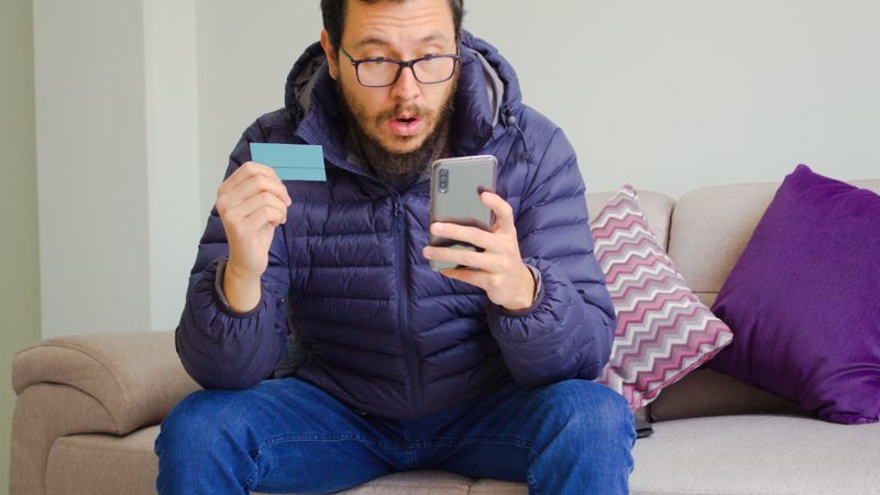 Imagem de um rapaz realizando compras online com seu cartão e smartphone nas mãos