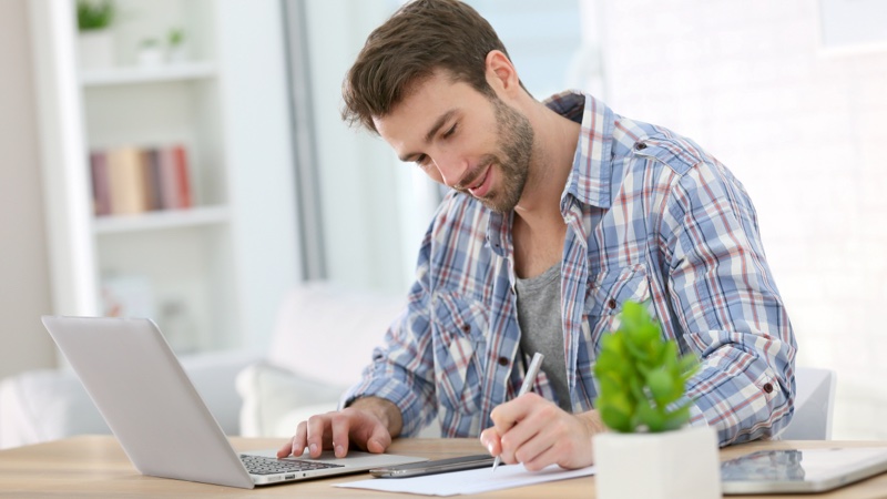 Homem usando notebook enquanto anota em folha de papel na mesa.