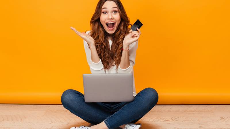 Mulher surpresa, sentada com notebook no colo e cartão na mão.