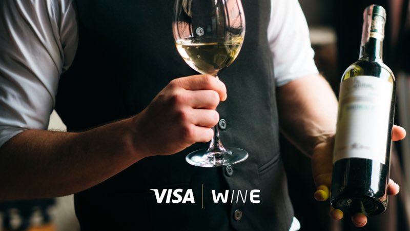 Wine Select Garçom com garrafa e taça de vinho em mãos, com os logotipos da Visa e Wine abaixo
