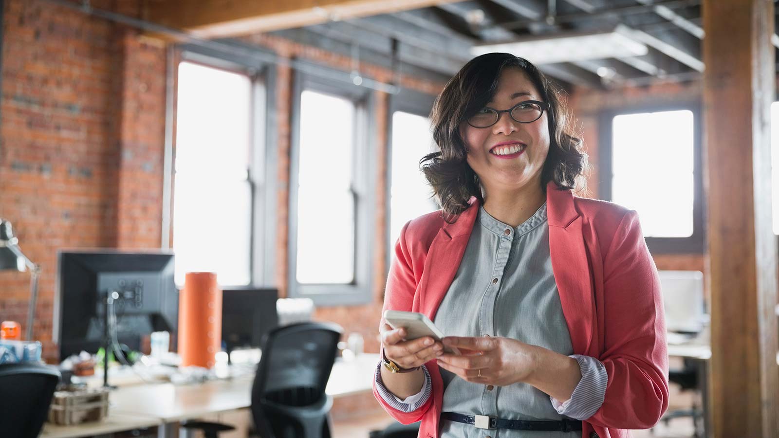 Woman with glasses holding phone in office setting.