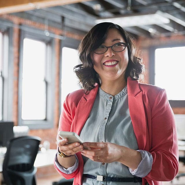 Woman with glasses  holding phone in office setting.